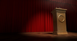 A regular theater stage with closed red curtains and a wooden debate podium lit by a single spotlight